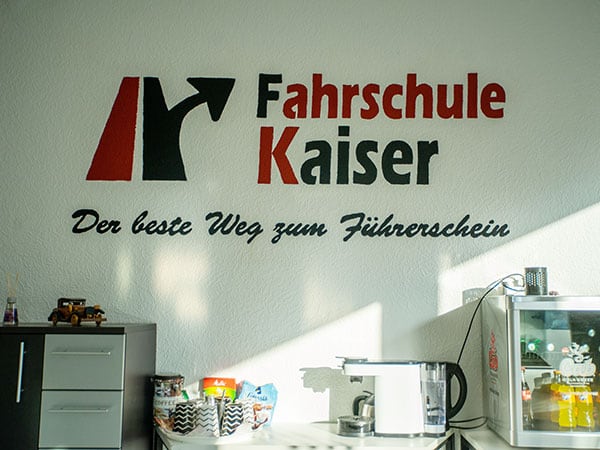 Die Erfrischungsecke der Fahrschule Kaiser: das Logo mit dem Schriftzug „Der beste Weg zum Führerschein“ ist auf die Wand gemalt, darunter stehen Wasserkocher, Kaffeemaschine, ein Korb mit Snacks und ein Getränkekühlschrank