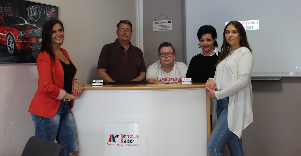 Fünf Mitarbeiterinnen und Mitarbeiter der Fahrschule Kaiser stehen und sitzen um einen weißen Empfangstresen, an dessen Front das Fahrschul-Logo klebt; links an der Wand ein gerahmtes Poster mit einem roten Sportwagen, rechts ein Whiteboard
