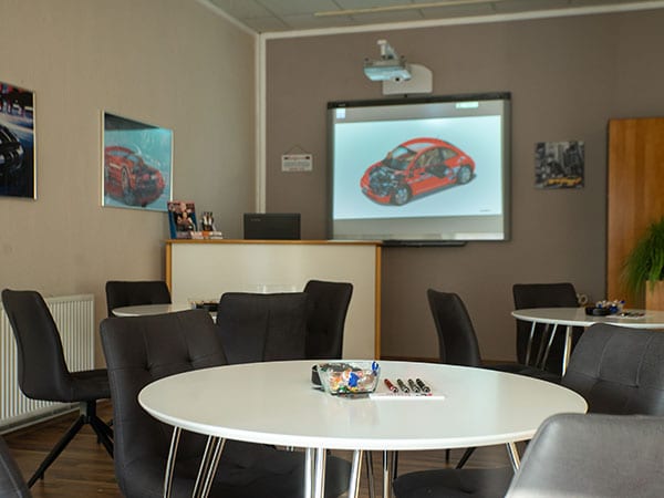 Theorieraum der Fahrschule Kaiser: weiße runde Tische mit schwarzen Stühlen vor einem an der Wand montierten Smartboard, auf dem die Schnittzeichnung eines roten Autos zu sehen ist